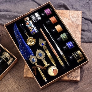 Op de afbeelding: Een kalligrafieset in een doos met kaartpatroon, bestaande uit een blauwe veer met gouden spikkels, twee metalen pennen, een waszegel met een blauwe steen, een briefopener, een glazen pot met wasparels, een smeltlepel en zes inktpotjes.