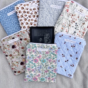 May include: A collection of seven quilted fabric pouches with various floral, cherry, and dotted patterns, each featuring a white Little Stitched Co. label. A black e-reader displaying the text Bourbon & Secrets is positioned in the centre.