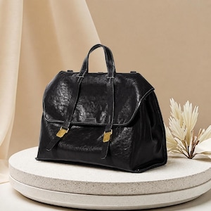 Pode incluir: Uma pasta de couro preta com acabamento texturizado, apresentando uma alça superior, uma aba dobrável e duas tiras verticais presas com fivelas de metal dourado. A bolsa tem uma base retangular estruturada e costuras visíveis nas bordas.