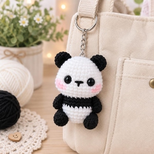 Puede incluir: Un pequeño llavero de panda hecho a mano en ganchillo, con cabeza y cuerpo blancos, orejas, brazos, piernas y sección media en negro. El panda tiene grandes ojos negros de plástico, una pequeña nariz bordada y mejillas de color rosa.