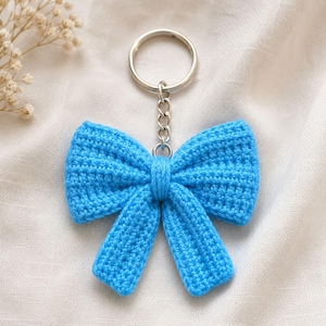 May include: A bright blue crochet bow keychain featuring a textured yarn construction with a central knot detail. The bow is attached to a silver-toned metal split ring by a short chain link, creating a functional accessory with a soft, fabric finish.