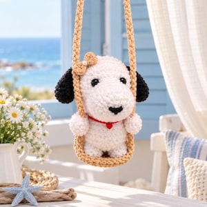 Puede incluir: Una figura de perro tejida a ganchillo en color blanco con orejas negras, un pequeño lazo beige en la cabeza y un collar rojo con un dije de corazón. El perro está sentado en un columpio tejido beige suspendido por dos cuerdas gruesas.