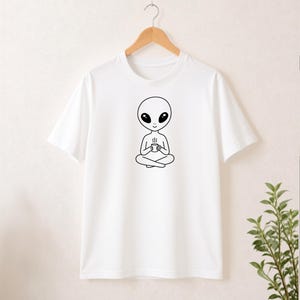 Könnte beinhalten: Ein weißes Kurzarm-T-Shirt mit einer schwarzen Strichgrafik eines Aliens, das im Schneidersitz sitzt und eine dampfende Tasse hält. Das Alien hat einen großen Kopf, ovale Augen und ein einfaches Lächeln, zentriert auf dem Brustbereich.