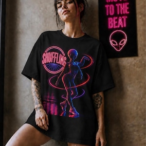 May include: A black t-shirt featuring a graphic print of an alien figure dancing in a neon-style aesthetic. The design includes the word SHUFFLING in a circular neon pink frame, with glowing red and blue light trails surrounding the alien character.