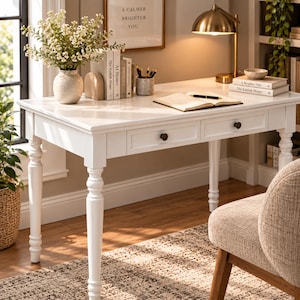 May include: A white wooden desk featuring four turned legs and two drawers with dark round knobs. The desk surface holds a white vase with white flowers, a stack of books, a pencil holder, and an open notebook with a black pen on top.
