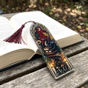 May include: A rectangular bookmark featuring a stained glass style illustration of a dragon perched on a stack of books, surrounded by candles, mushrooms, and foliage, topped with a deep red tassel and set against an open hardcover book.