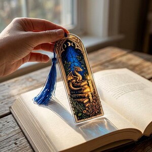 May include: A rectangular bookmark with a rounded top featuring a stained glass style illustration of a golden dragon perched on a stack of books, surrounded by foliage and candles, finished with a blue tassel attached to the top center.