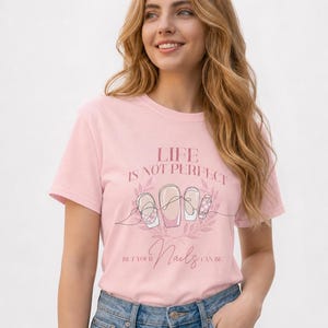 May include: A light pink short-sleeve t-shirt featuring a graphic print with the text "LIFE IS NOT PERFECT BUT YOUR NAILS CAN BE" and an illustration of four manicured fingernails surrounded by a delicate leaf wreath in shades of pink.