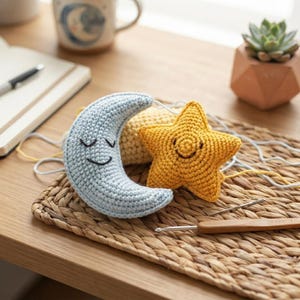 Peut inclure: Un croissant de lune bleu clair et une étoile jaune, tous deux réalisés au crochet, présentent des expressions faciales brodées. La lune a les yeux fermés et sourit, tandis que l'étoile affiche un large sourire. Du fil blanc et un crochet sont présents.