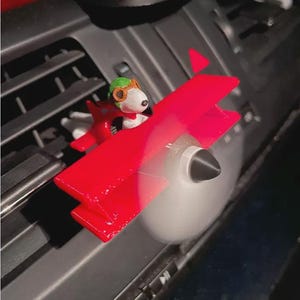 May include: A red biplane car air freshener clip featuring a white cartoon dog character wearing a green aviator cap with brown goggles. The airplane has a black nose cone and a spinning propeller, designed to attach to a vehicle dashboard air vent.