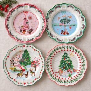 Puede incluir: Cuatro platos redondos de cerámica con bordes festoneados, cada uno con una ilustración navideña única. Los diseños incluyen un árbol rosa, un paisaje invernal con ciervos, una escena de pueblo nevado y un árbol decorado con regalos.