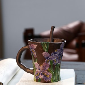 Può includere: Una tazza in ceramica marrone con superficie testurizzata decorata con fiori di iris viola e steli verdi dipinti a mano. La tazza presenta un manico curvo e un cucchiaino in ceramica marrone abbinato all'interno con dettagli floreali.