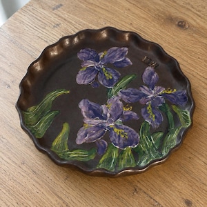 Peut inclure: Une assiette ronde en céramique brun foncé avec un bord festonné. La surface présente un motif peint à la main de trois fleurs d'iris violettes avec des feuilles et des tiges vertes. Les pétales ont des coups de pinceau et des touches jaunes.