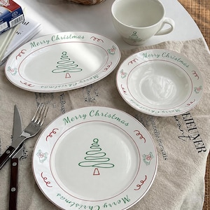 Puede incluir: Un juego de vajilla de cerámica blanca con un árbol de Navidad dibujado en líneas verdes, texto Merry Christmas en rojo y verde, y pequeñas ilustraciones de campanas. El set incluye una fuente ovalada, un plato, un cuenco y una taza.
