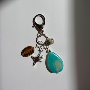 May include: A silver-toned metal keychain featuring a lobster clasp and a circular ring. Attached are four charms: a brown tiger's eye oval bead, a silver four-pointed star, a small speckled beige bead, and a large, smooth, teardrop-shaped turquoise stone.