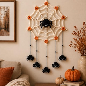 May include: A cream-colored crocheted spider web wall hanging featuring small orange flowers at the intersections and a central black crocheted spider. Five additional black crocheted spiders hang from the web on thin black cords with beads.