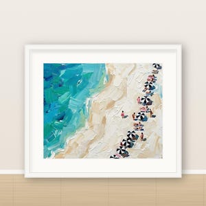 May include: An oil painting in a white frame depicting an aerial view of a beach with thick, textured brushstrokes. The scene features vibrant turquoise and blue ocean waves meeting a sandy beige shore with small black and white umbrellas.