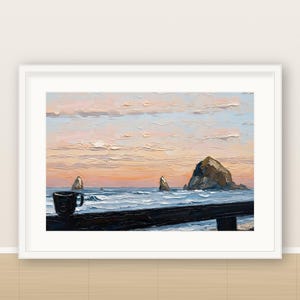 May include: An oil painting in a white frame depicting a dark mug resting on a dark railing overlooking a turbulent ocean at sunset. Large rock formations rise from the water under a sky filled with soft pink, orange, and blue clouds.