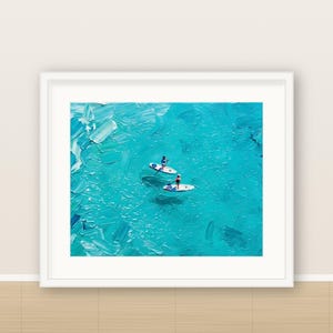 May include: A framed oil painting featuring two individuals on white stand-up paddleboards in vibrant turquoise water. The artwork displays thick, textured brushstrokes that capture the movement and light of the ocean surface.