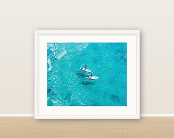 May include: A framed oil painting featuring two individuals on white stand-up paddleboards in vibrant turquoise water. The artwork displays thick, textured brushstrokes that capture the movement and light of the ocean surface.