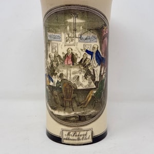 May include: A cream-colored ceramic vase with a black rim and base. The front features a detailed, oval-shaped illustration of a group of men gathered around a table, with the text "Mr. Pickwick addresses the Club" printed on a small plaque below.