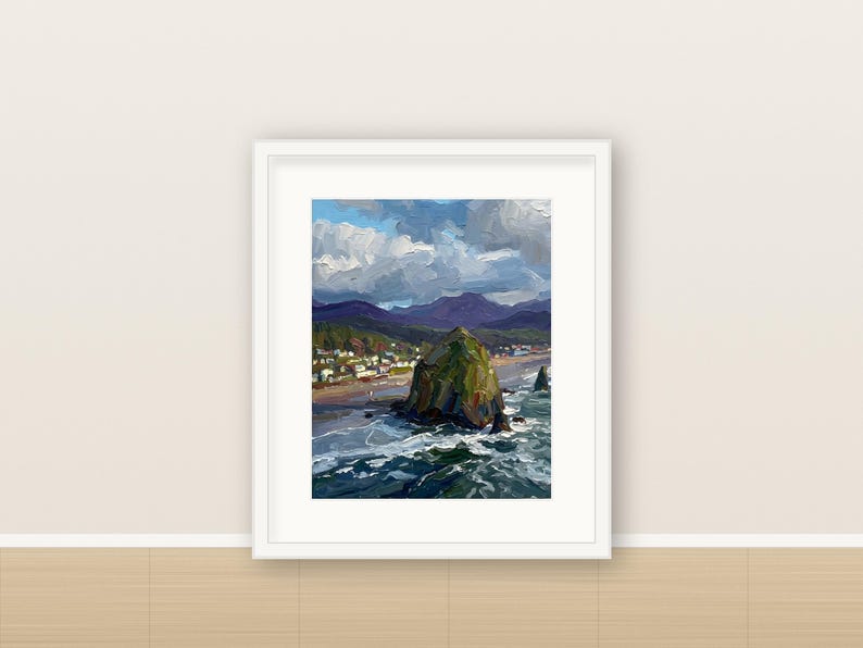 May include: A framed oil painting depicting a coastal landscape with a large green rock formation in the ocean, a sandy beach with small buildings, and dark mountains under a cloudy blue sky. The artwork is enclosed in a white frame.