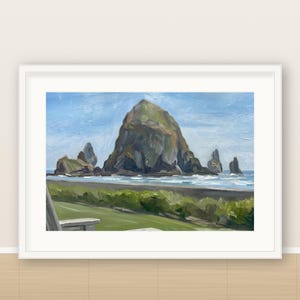 May include: A framed oil painting depicting a large, dark, jagged rock formation rising from the ocean near a sandy beach. The foreground features green coastal vegetation and a partial view of a wooden chair, all under a bright blue sky.