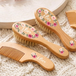 Puede incluir: Dos cepillos de madera con cerdas suaves y dos peines de madera, todos con un diseño de corona floral en rosa y blanco con el nombre Olive impreso en letra cursiva blanca. Los artículos están dispuestos sobre una superficie blanca texturizada.