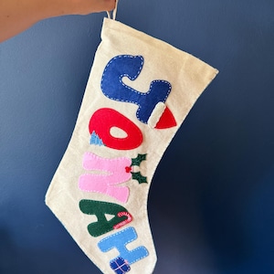 May include: A beige fabric Christmas stocking featuring the name JONAH appliqued in colourful felt letters. The J is blue with a red hat, O is red, N is pink with holly leaves, A is dark green with a candy cane, and H is light blue with a gift.