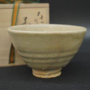 May include: A beige ceramic tea bowl with a subtle crackle glaze finish and horizontal ribbed detailing along the exterior walls. The bowl features a flared rim and a sturdy footed base, presented alongside a wooden storage box with ribbon.