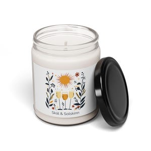 May include: A white candle jar with a black lid. The label features a sun, four glasses of wine, and the text "Skål & Solskinn".