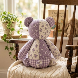 May include: A stuffed bear toy featuring a patchwork design of lavender and white fabrics. The bear is constructed from alternating panels of floral patterns and a classic gingham check print, with rounded ears and a small tail on the back.