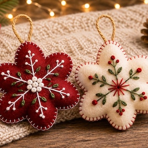 Peut inclure: Deux décorations en feutrine en forme d'étoile avec des boucles de suspension. L'une est rouge foncé avec des motifs de flocons de neige brodés en blanc et des perles blanches au centre. L'autre est crème avec des brins verts, des baies rouges.