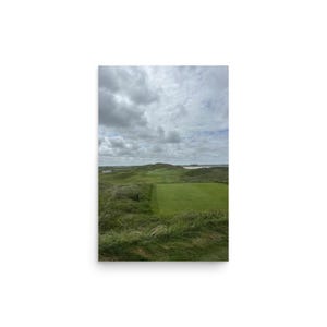 May include: A landscape view of a green golf course fairway surrounded by rolling grassy dunes and hills under a cloudy, overcast sky. The scene features patches of tall wild grass in the foreground and a distant horizon line with water.
