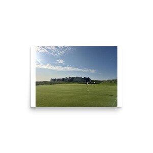 May include: A wide view of a green golf course putting surface with a flagstick in the foreground. In the distance, a row of multi-storey buildings sits against a clear blue sky with light clouds. The grass is short and manicured across the field.