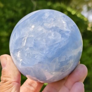 May include: A polished spherical stone in a pale sky blue colour with translucent and opaque white cloudy inclusions throughout its surface. The orb displays a smooth, reflective texture with visible internal crystalline layers and natural mineral patterns.