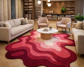 May include: An irregular, organic-shaped area rug featuring a gradient of pink and red wavy bands. The rug is positioned in a room with two light-colored sofas, two armchairs, a round white coffee table, and a gold multi-globe chandelier.