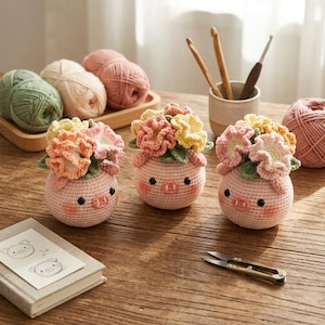 May include: Three round, light pink crocheted pig figures, each topped with a cluster of crocheted flowers in shades of yellow, peach, and pink. The pigs feature small black eyes and embroidered snouts. Nearby are yarn balls, crochet hooks, and a notebook.