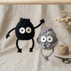 May include: Two crocheted ornaments hanging from a wooden rod. One is a black round figure with thin arms and legs and two large white eyes with black pupils. The other is a grey round figure with similar eyes and dangling arms and legs.