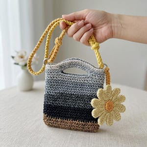 May include: A small, rectangular crochet handbag featuring a horizontal ombre pattern that transitions from light gray to dark gray, black, and brown. The bag has a yellow crocheted shoulder strap and a large, yellow crocheted flower charm attached.