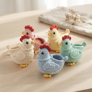 May include: A set of four small, hollow, crocheted chicken-shaped containers in pastel shades of cream, pink, yellow, and light blue. Each chicken features a red comb, a yellow beak, and black bead eyes, with a hollow center for holding small items.