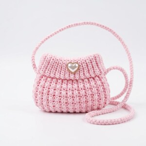 May include: A light pink crocheted handbag featuring a textured knit pattern, a single long shoulder strap, and a central decorative heart-shaped embellishment with a white faux pearl center set in a gold-toned metal frame on the front flap.