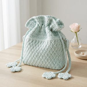 May include: A light mint green crochet drawstring bag with a textured stitch pattern. The bag features a gathered top closure and two long straps, each ending in a small, four-petaled crochet flower detail. The design is soft and uniform in color.