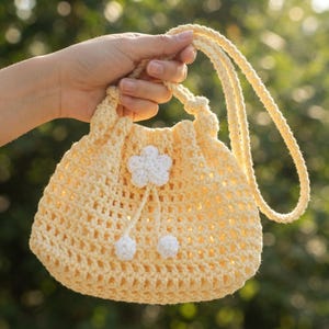 May include: A pale yellow crochet drawstring bag featuring an open-weave stitch pattern, a long shoulder strap, and a decorative white crocheted flower centered on the front with two small white pom-poms hanging from the flower by thin cords.