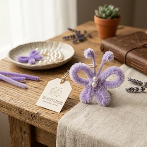 May include: A light purple pipe cleaner butterfly ornament featuring two small white pearl beads at the center. A beige paper tag is attached by a twine string, printed with the text THE FUZZY GARDEN Handcrafted with Care and a butterfly logo.