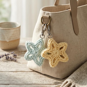 May include: A beige linen tote bag featuring two crochet star-shaped charms attached to a metal D-ring with silver-toned lobster clasps. One star is light blue and the other is pale yellow, both showcasing an openwork, five-pointed design.