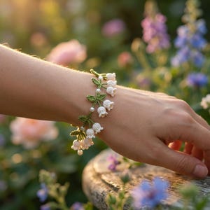 May include: A crochet bracelet featuring white lily of the valley flowers with green stems and leaves, accented by small white beads. The floral design is crafted with fine thread, showcasing delicate bell-shaped blossoms arranged along the band.