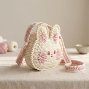 May include: A round, cream-colored crochet bag shaped like a rabbit face. The bag features two upright ears, pink accents on the ears and cheeks, and embroidered pink eyes and a small nose. It includes a matching cream and pink crochet shoulder strap.