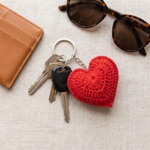 May include: A red crocheted heart keyring attached to a silver metal ring with two silver keys and one black plastic key. The keyring is positioned near a tan leather wallet and a pair of brown tortoiseshell sunglasses with dark lenses.