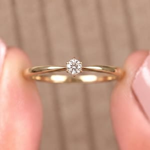 以下が含まれることがあります： 6本の爪で留められた小さなラウンドカットの透明な宝石が特徴のゴールドリング。バンドは滑らかで光沢があり、中央の石に向かってわずかに細くなっており、繊細で洗練された外観のジュエリーに仕上がっています。
